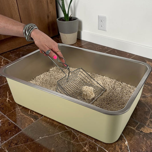 Person using a scoop to clean a large metal cat litter box on a tiled floor.
