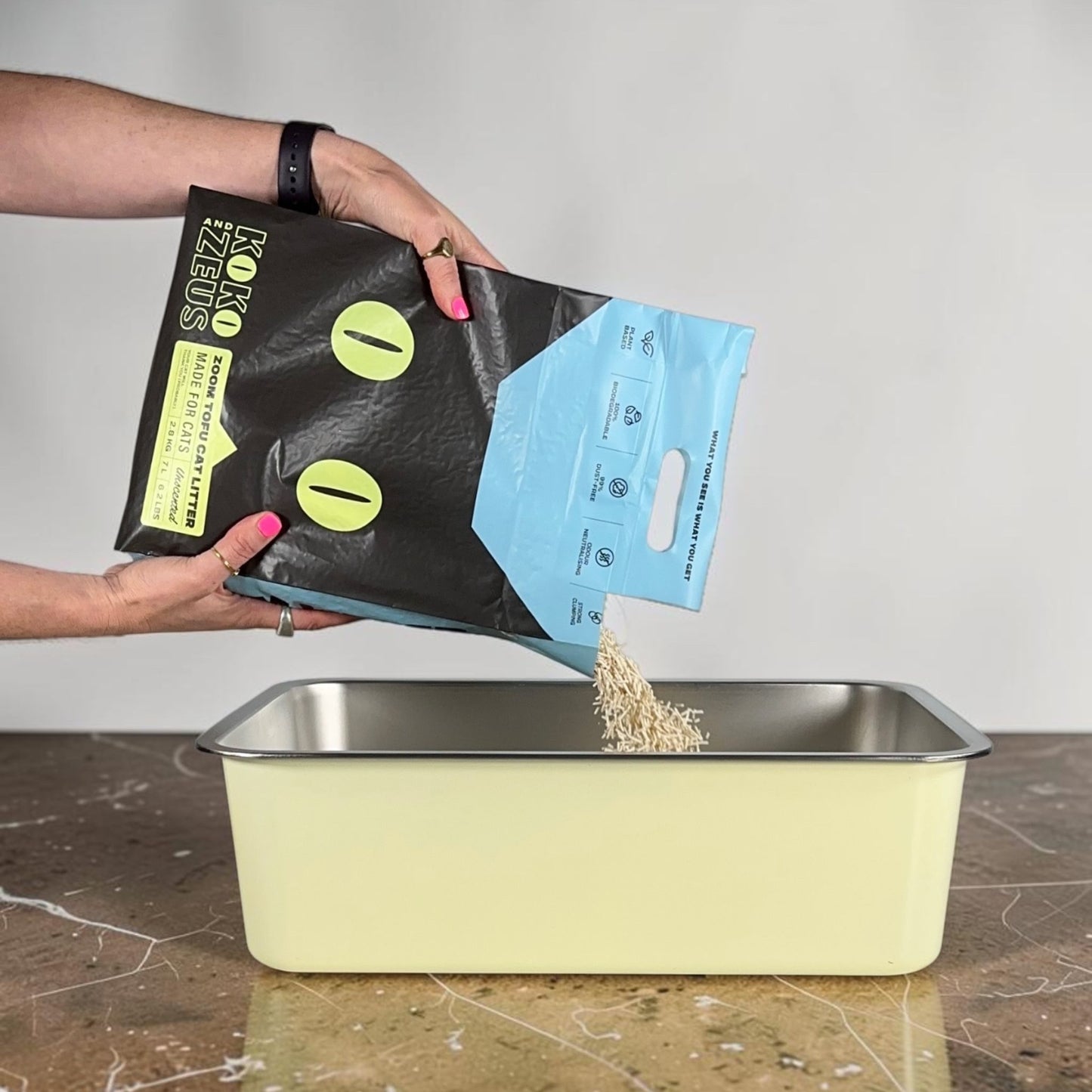 Person pouring ZOOM Tofu Cat Litter into a stainless steel litter box on a brown marble surface.